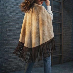 May include: A tan and brown patterned poncho featuring a textured surface and long dark brown fringe along the bottom hem. The garment is worn over a white long-sleeved shirt and blue denim jeans, paired with dark brown high-heeled shoes.