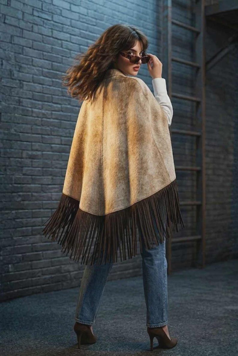 May include: A tan and brown patterned poncho featuring a textured surface and long dark brown fringe along the bottom hem. The garment is worn over a white long-sleeved shirt and blue denim jeans, paired with dark brown high-heeled shoes.