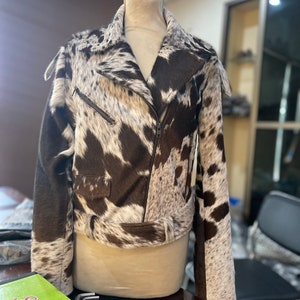 May include: A brown and white cowhide leather jacket with a zipper closure and a belt at the waist.
