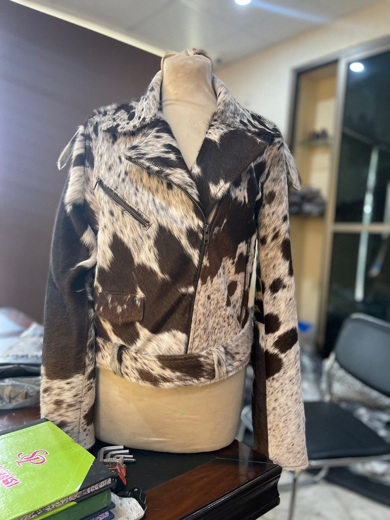 May include: A brown and white cowhide leather jacket with a zipper closure and a belt at the waist.