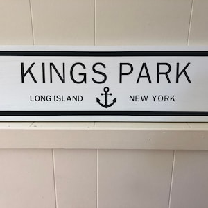 Puede incluir: Un letrero de madera blanco con letras negras y un símbolo de ancla negro. El letrero dice "Kings Park, Long Island, New York".