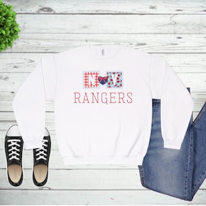 May include: White sweatshirt with the text "RANGERS" in red script. Above the text are two decorative squares with "NY" inside. The "N" is in a red and white checkered square, and the "Y" is in a blue and red floral square. Black sneakers and blue jeans are also in the image.