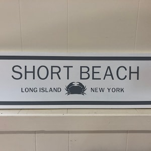 Puede incluir: Un letrero de madera blanco con un borde gris y texto que dice "SHORT BEACH LONG ISLAND NEW YORK". Una silueta de cangrejo gris está centrada entre las palabras "LONG ISLAND" y "NEW YORK".