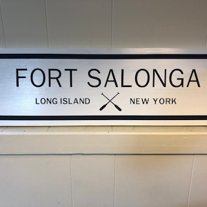 Puede incluir: Un letrero plateado con letras negras y un borde negro que dice "FORT SALONGA LONG ISLAND NEW YORK" con un par de remos cruzados entre las palabras "LONG ISLAND" y "NEW YORK".