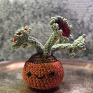May include: A handmade crochet plant with a smiling face. The orange pot has a brown top and holds three green and red plant heads. The plant has a friendly, whimsical design, perfect for home decor or as a unique gift.