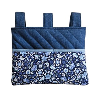 May include: A rectangular fabric hanging organizer featuring a dark blue quilted upper section with three vertical straps at the top. The lower section is a pocket with a dark blue background, white floral patterns, and small bird motifs.