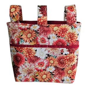 Könnte beinhalten: Eine Stofftasche mit einem lebendigen Blumenmuster aus orangefarbenen, roten, gelben und weißen Chrysanthemen. Die Tasche verfügt über drei vertikale Stoffriemen an der Oberseite sowie eine horizontale Tasche auf der Vorderseite.
