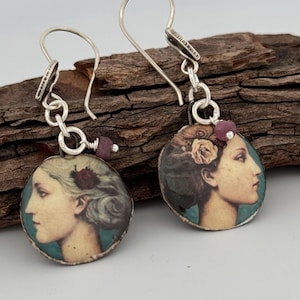 May include: A pair of silver earrings with a round pendant featuring a vintage illustration of a woman's face with a flower in her hair. The pendants are accented with small pink gemstones.