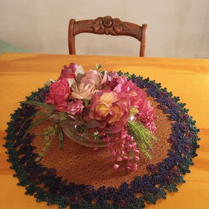 May include: A floral arrangement in a glass bowl, with pink and yellow roses, rests on a brown oval placemat with a dark blue and green beaded trim. A wooden chair with a rose carving is in the background.