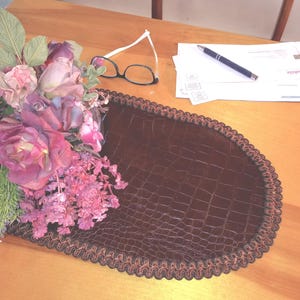 May include: A brown oval placemat with a textured, crocodile-like pattern and a decorative trim. A bouquet of pink and purple flowers, eyeglasses, and a pen rest on a wooden surface.