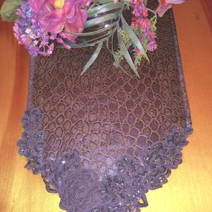 May include: A dark brown table runner with a textured, crocodile-like pattern. The runner is adorned with a decorative, dark purple floral lace trim. A bouquet of purple and pink flowers sits atop the runner.