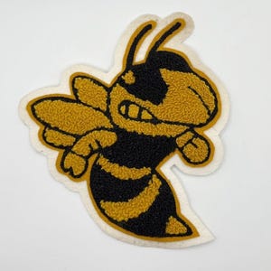 May include: A yellow and black embroidered bee patch with a white border. The bee has a smiling face and is in a flying position. The patch is likely for a jacket or bag.