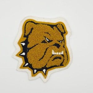 May include: A patch featuring a detailed, textured bulldog head in shades of gold and brown, outlined in black with white accents. The patch has a white border with black and white spikes.