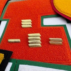 Puede incluir: Primer plano de una letra de fieltro rojo con barras doradas. La letra está delineada en verde y blanco. El fieltro tiene una superficie texturizada.