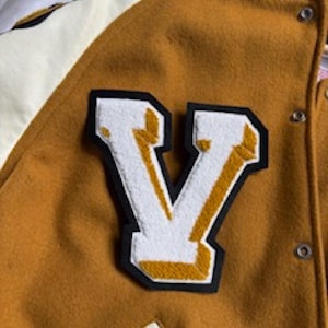 May include: Mustard yellow varsity jacket with a large white and gold letter "V" patch. The jacket has a button-up front and a cream-colored lining. The letter "V" is outlined in black.