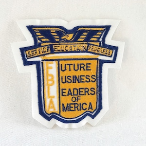 Puede incluir: Parche bordado con el texto "SERVICE EDUCATION PROGRESS" y "FUTURE BUSINESS LEADERS OF AMERICA". El parche tiene forma de escudo con borde azul y letras doradas y azules. Un águila en la parte superior.
