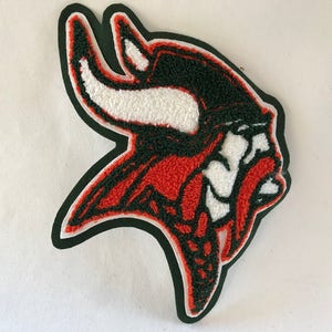 May include: A red, black, and white embroidered patch featuring a stylized horned head. The patch has a black border and a textured, raised appearance. The design is reminiscent of a mascot or emblem.