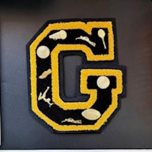 May include: A black and gold letter "G" patch with a yellow border and black interior. The letter is decorated with gold-colored sports-themed embellishments, including golf balls and golf clubs. The patch is likely for a varsity jacket or similar apparel.