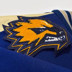 Könnte beinhalten: Nahaufnahme einer blauen Jacke mit einem großen, gestickten Aufnäher. Der Aufnäher zeigt einen stilisierten Wolfskopf in Gelb und Marineblau mit weißen Details. Die Jacke hat einen cremefarbenen Lederkragen und gestreifte marine- und cremefarbene Manschetten.