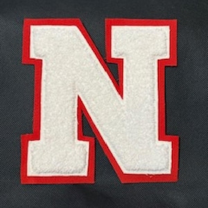 May include: A white felt letter "N" with a red border, set against a dark gray textured background. The letter is thick and blocky, with a classic collegiate style. Ideal for sports apparel or crafts.