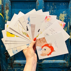 Puede incluir: Una mano sostiene un abanico de tarjetas de felicitación ilustradas sobre un fondo azul. Las tarjetas presentan ilustraciones en acuarela de animales, flores y texto. El texto incluye frases como "Might spirit stones at tod" y "my work is dubious and frivolous."