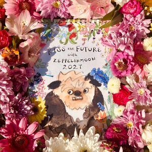 May include: A spiral-bound calendar featuring a whimsical illustration of a furry creature with prominent teeth, surrounded by a vibrant border of fresh pink, yellow, white, and red flowers. The text reads KISS the FUTURE with ZEPPELINMOON 2027.