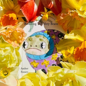 Peut inclure: Illustration colorée d'une grenouille vêtue d'une tenue violette ornée d'étoiles jaunes, entourée de fleurs jaunes et oranges. Le texte "Rainfrog The Very Brave" et "Amber Fossey" est également visible.