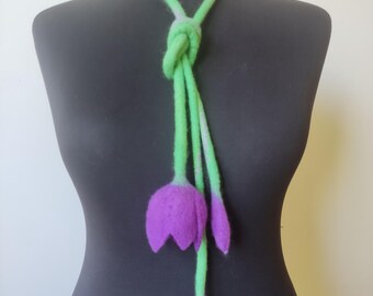 Puede incluir: Un collar de fieltro hecho a mano con un cordón verde anudado que se divide en tres hebras. Las hebras terminan con una flor morada en forma de tulipán y dos capullos. El collar se exhibe en un maniquí negro.
