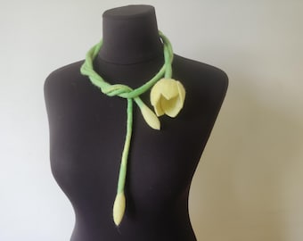 Può includere: Una collana fatta a mano in feltro con un cordoncino ritorto verde chiaro, un fiore di tulipano giallo e boccioli. La collana è esposta su un manichino nero, mettendo in risalto il design floreale e l'artigianato.