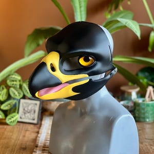 Puede incluir: Una máscara de pájaro negra, amarilla y blanca con ojos amarillos grandes y expresivos y una lengua rosa. La máscara se exhibe en una cabeza de maniquí gris. La máscara está hecha de un material duro y tiene un diseño realista.