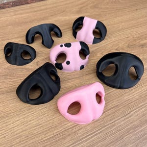 May include: A collection of seven three-dimensional animal nose shapes in various colours and patterns. The set includes solid black noses, solid pink noses, and one pink nose with black spots, all featuring two distinct nostril openings.