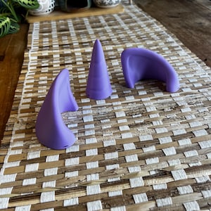 K&ouml;nnte beinhalten: Ein Set aus drei violetten Silikon-Accessoires, bestehend aus zwei geschwungenen, ohrf&ouml;rmigen Teilen und einem zentralen, kegelf&ouml;rmigen Horn. Jeder Gegenstand weist eine glatte, matte Oberfl&auml;che und eine einheitliche violette Farbe auf.