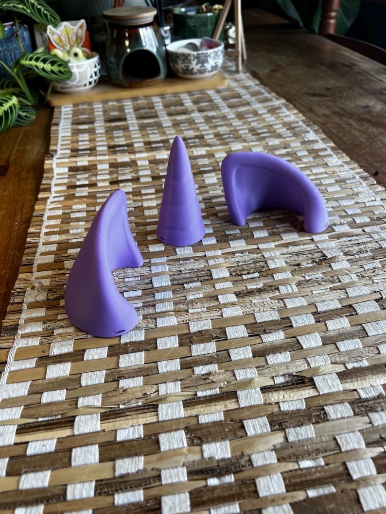 K&ouml;nnte beinhalten: Ein Set aus drei violetten Silikon-Accessoires, bestehend aus zwei geschwungenen, ohrf&ouml;rmigen Teilen und einem zentralen, kegelf&ouml;rmigen Horn. Jeder Gegenstand weist eine glatte, matte Oberfl&auml;che und eine einheitliche violette Farbe auf.