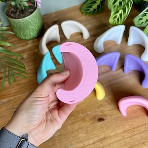 K&ouml;nnte beinhalten: Eine Sammlung kleiner, halbmondf&ouml;rmiger Ohr-Aufs&auml;tze aus einfarbigem Kunststoff in verschiedenen Pastellt&ouml;nen wie Rosa, Blau, Lila, Gelb und Wei&szlig;. Jedes Teil verf&uuml;gt &uuml;ber zwei kleine kreisf&ouml;rmige L&ouml;cher an den oberen und unteren Kanten.