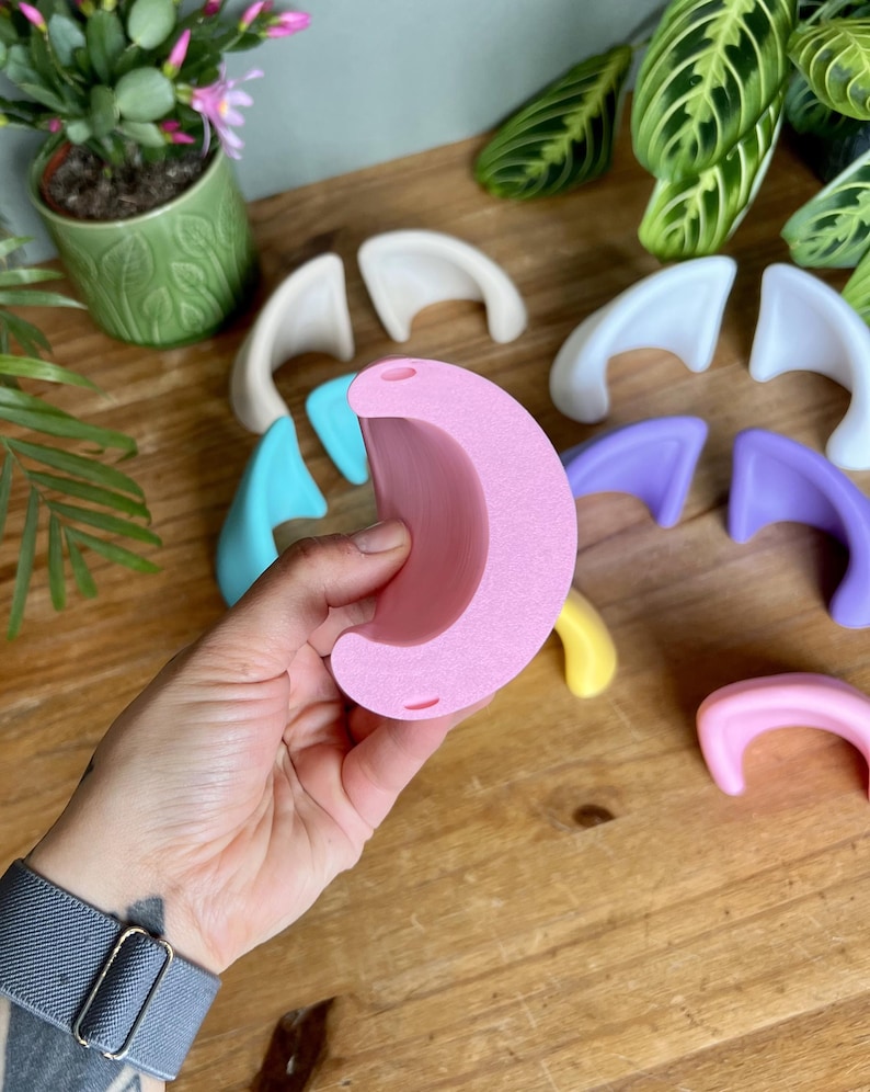 K&ouml;nnte beinhalten: Eine Sammlung kleiner, halbmondf&ouml;rmiger Ohr-Aufs&auml;tze aus einfarbigem Kunststoff in verschiedenen Pastellt&ouml;nen wie Rosa, Blau, Lila, Gelb und Wei&szlig;. Jedes Teil verf&uuml;gt &uuml;ber zwei kleine kreisf&ouml;rmige L&ouml;cher an den oberen und unteren Kanten.