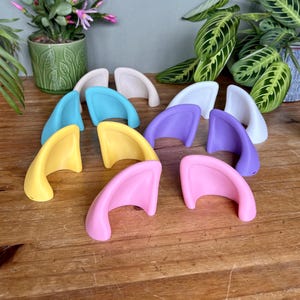 May include: A collection of ten small, curved, ear-shaped accessories in various solid colours including light blue, yellow, pink, purple, white, and beige. Each piece features a smooth, rounded design with a hollow underside, arranged in pairs.