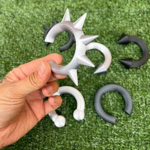 Pode incluir: Uma cole&ccedil;&atilde;o de braceletes em forma de C com espinhos pontiagudos, dispon&iacute;veis em preto s&oacute;lido, branco s&oacute;lido e um acabamento prateado. Estes acess&oacute;rios circulares t&ecirc;m uma abertura e uma superf&iacute;cie lisa com textura mate.