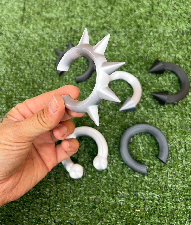 Pode incluir: Uma cole&ccedil;&atilde;o de braceletes em forma de C com espinhos pontiagudos, dispon&iacute;veis em preto s&oacute;lido, branco s&oacute;lido e um acabamento prateado. Estes acess&oacute;rios circulares t&ecirc;m uma abertura e uma superf&iacute;cie lisa com textura mate.