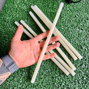 Puede incluir: Varios barras de pegamento blanquecinas están dispuestas sobre una superficie verde. Los palos cilíndricos se sostienen en una mano, algunos se superponen. El fondo incluye un objeto negro y una correa gris.