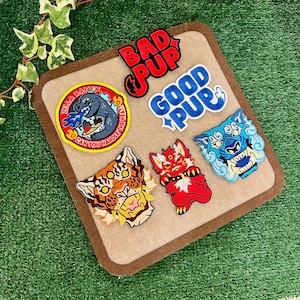 Puede incluir: Una colección de parches bordados con varios diseños. Incluye un monstruo tipo Godzilla con el texto "Zilla Ramen" y "Can you handle the heat?", un parche rojo "Bad Pup" y un parche azul "Good Pup". Otros parches representan caras de tigre y criaturas míticas.