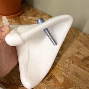 Pode incluir: Um acess&oacute;rio em forma de orelha de espuma branca triangular, com um brinco de pino met&aacute;lico azul-claro liso com uma haste longa e c&oacute;nica. O brinco est&aacute; inserido atrav&eacute;s da parte superior da estrutura branca, curva e em forma de orelha.