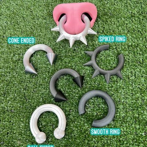 Pode incluir: Uma cole&ccedil;&atilde;o de joias corporais incluindo um modelo de nariz rosa com um anel prateado de espinhos, uma ferradura prateada com pontas c&oacute;nicas, um anel preto de espinhos, uma ferradura preta com pontas c&oacute;nicas, um anel cinzento liso.