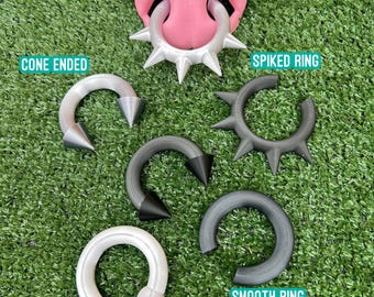 May include: A collection of circular body jewelry including a silver spiked ring on a pink nose model, a white cone-ended horseshoe, a black spiked ring, a black cone-ended horseshoe, a smooth grey ring, and a white ball-ended horseshoe ring.