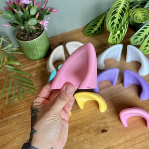 K&ouml;nnte beinhalten: Eine Sammlung glatter, geschwungener, ohrf&ouml;rmiger Accessoires in verschiedenen Unifarben wie Pink, Gelb, Lila, Blau und Wei&szlig;. Jedes Teil hat eine abgerundete, dreieckige Form mit einer ausgeh&ouml;hlten Mitte und ist zur Befestigung gedacht.