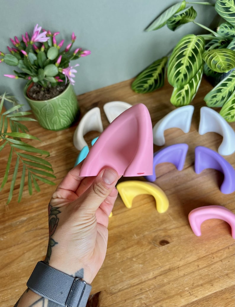 K&ouml;nnte beinhalten: Eine Sammlung glatter, geschwungener, ohrf&ouml;rmiger Accessoires in verschiedenen Unifarben wie Pink, Gelb, Lila, Blau und Wei&szlig;. Jedes Teil hat eine abgerundete, dreieckige Form mit einer ausgeh&ouml;hlten Mitte und ist zur Befestigung gedacht.