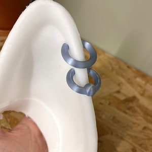 Pode incluir: Dois brincos de argola circulares em cinzento mate est&atilde;o presos &agrave; borda de um expositor branco curvo em forma de orelha. As argolas t&ecirc;m um acabamento liso e s&oacute;lido e est&atilde;o posicionadas uma sobre a outra ao longo da borda exterior.