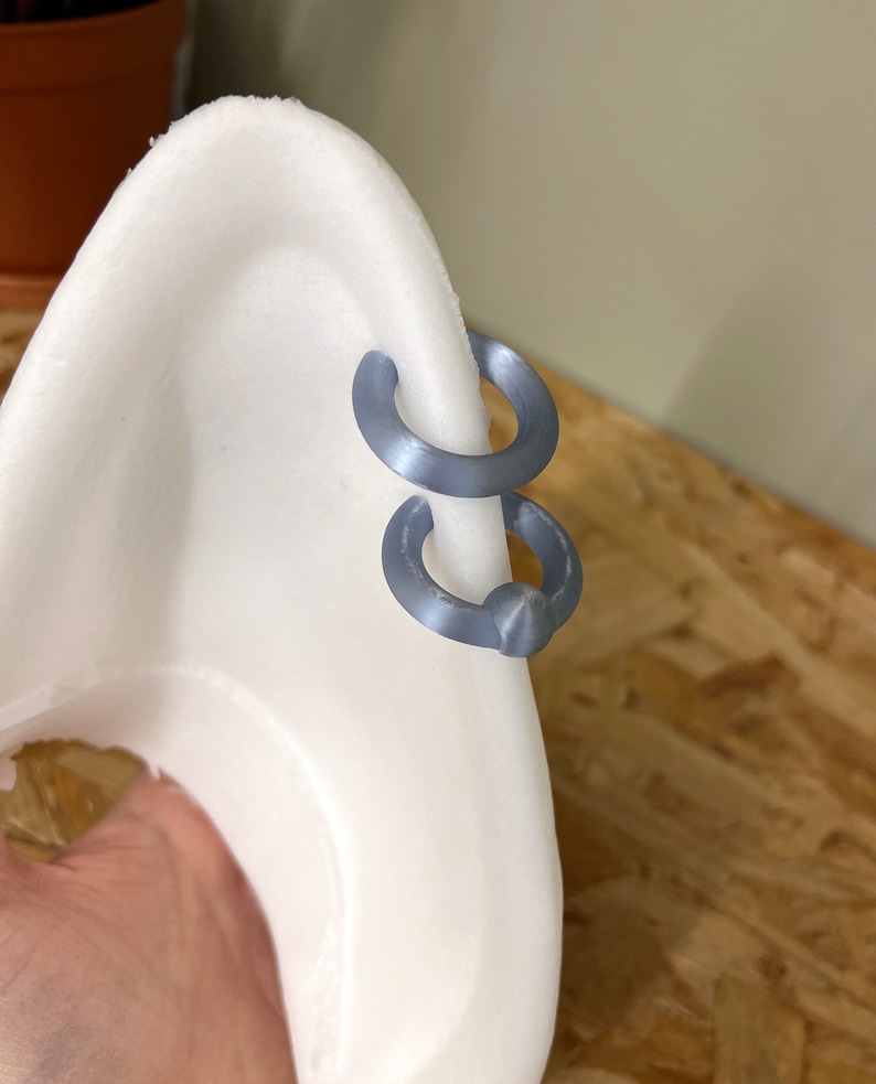 Pode incluir: Dois brincos de argola circulares em cinzento mate est&atilde;o presos &agrave; borda de um expositor branco curvo em forma de orelha. As argolas t&ecirc;m um acabamento liso e s&oacute;lido e est&atilde;o posicionadas uma sobre a outra ao longo da borda exterior.