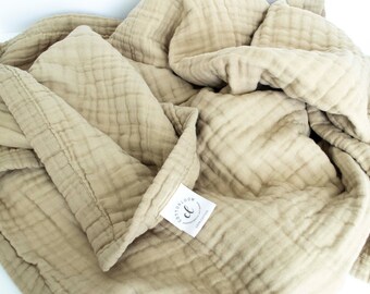 May include: A beige cotton muslin throw blanket with a stitched pattern. The blanket is folded and has a white label with the text "Cotton Cloud" and a stylized "C" logo.