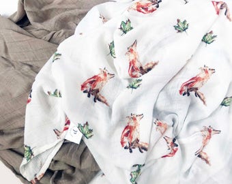 May include: White muslin swaddle blanket with a repeating pattern of orange foxes and green leaves. The blanket is folded and partially draped over a brown muslin blanket.