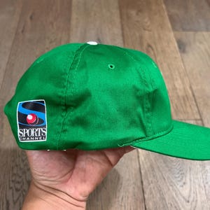 May include: A bright green baseball cap with a curved brim. The cap features a patch on the side with the text "SPORTS CHANNEL" and a logo with blue, red, and white elements. The cap is made of a smooth fabric.